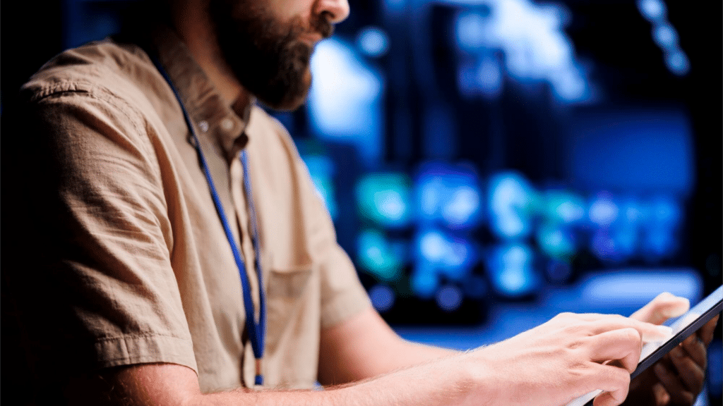 A imagem mostra um homem de perfil, usando uma camisa clara e um crachá de identificação, enquanto interage com um tablet. O ambiente ao fundo é digitalmente sofisticado, com imagens em tons de azul e ícones, possivelmente representando gráficos ou dados, sugerindo que ele está realizando uma tarefa técnica ou de auditoria em um ambiente digital ou de controle de informações. O foco está na tela do tablet e no gesto do homem, que parece estar analisando ou operando dados.