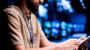 A imagem mostra um homem de perfil, usando uma camisa clara e um crachá de identificação, enquanto interage com um tablet. O ambiente ao fundo é digitalmente sofisticado, com imagens em tons de azul e ícones, possivelmente representando gráficos ou dados, sugerindo que ele está realizando uma tarefa técnica ou de auditoria em um ambiente digital ou de controle de informações. O foco está na tela do tablet e no gesto do homem, que parece estar analisando ou operando dados.