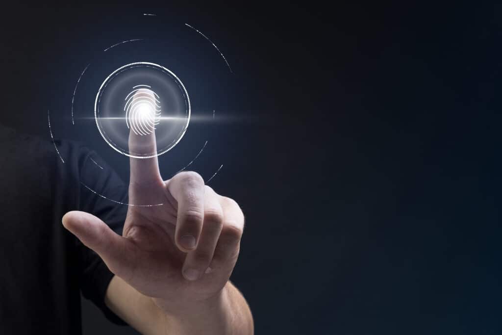 A imagem mostra uma pessoa utilizando um sistema de autenticação por impressão digital, com um dedo tocando um sensor de digital futurista. O sensor é visualizado em um formato digitalizado, com uma impressão digital luminosa no centro, cercada por círculos e linhas que indicam o processo de escaneamento. O fundo é escuro, destacando a tecnologia de segurança digital avançada.