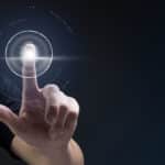 A imagem mostra uma pessoa utilizando um sistema de autenticação por impressão digital, com um dedo tocando um sensor de digital futurista. O sensor é visualizado em um formato digitalizado, com uma impressão digital luminosa no centro, cercada por círculos e linhas que indicam o processo de escaneamento. O fundo é escuro, destacando a tecnologia de segurança digital avançada.