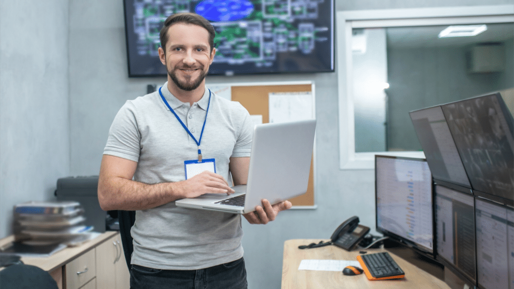 A imagem mostra um profissional sorridente segurando um laptop em uma sala de monitoramento. Ele está vestindo uma camiseta cinza e um crachá de identificação. Ao fundo, há várias telas de computador, que exibem gráficos e imagens de câmeras de segurança. A sala parece ser um centro de controle, com equipamentos de monitoramento e uma decoração simples, com paredes neutras e um telefone fixo sobre a mesa. O ambiente transmite a ideia de um local dedicado à vigilância e segurança eletrônica.