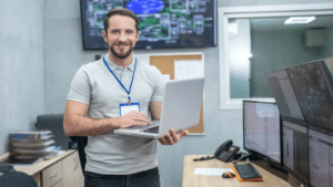 A imagem mostra um profissional sorridente segurando um laptop em uma sala de monitoramento. Ele está vestindo uma camiseta cinza e um crachá de identificação. Ao fundo, há várias telas de computador, que exibem gráficos e imagens de câmeras de segurança. A sala parece ser um centro de controle, com equipamentos de monitoramento e uma decoração simples, com paredes neutras e um telefone fixo sobre a mesa. O ambiente transmite a ideia de um local dedicado à vigilância e segurança eletrônica.