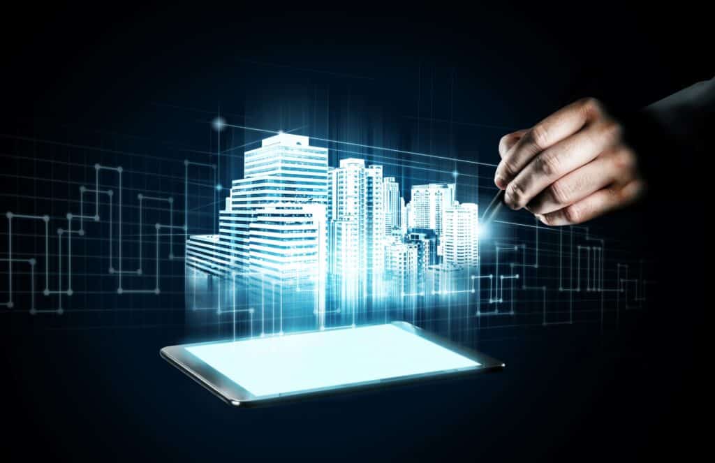 A imagem apresenta um conceito futurista de automação predial e cidades inteligentes. Um tablet emite uma projeção holográfica de um conjunto de edifícios, enquanto uma mão com uma caneta digital interage com a projeção, como se estivesse desenhando ou programando os prédios. O fundo escuro, com circuitos e gráficos digitais, reforça a ideia de tecnologia avançada, controle remoto e integração de sistemas para gestão de edifícios inteligentes, como iluminação, segurança, energia e climatização.