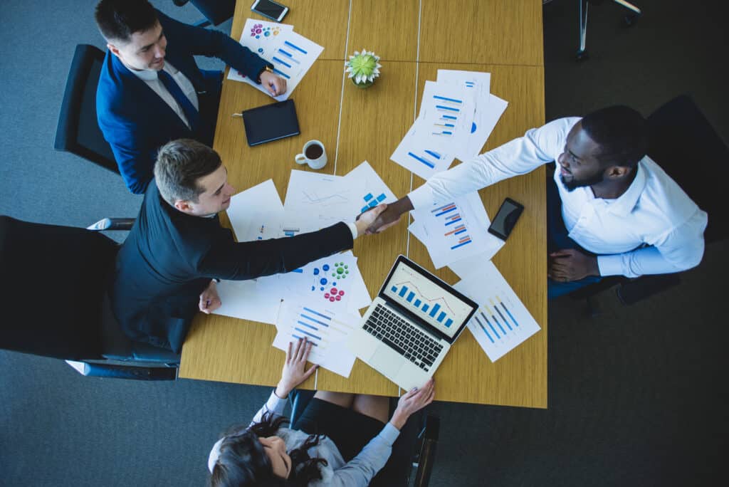 A imagem mostra quatro profissionais reunidos ao redor de uma mesa de reuniões, coberta por relatórios impressos com gráficos coloridos, planilhas e um notebook aberto exibindo dados financeiros. Dois deles estão apertando as mãos, indicando o fechamento de um acordo ou parceria. O ambiente é corporativo e bem iluminado, transmitindo uma atmosfera de colaboração, estratégia e tomada de decisão, associada a reuniões de negócios, análise de desempenho e gestão de riscos corporativos.