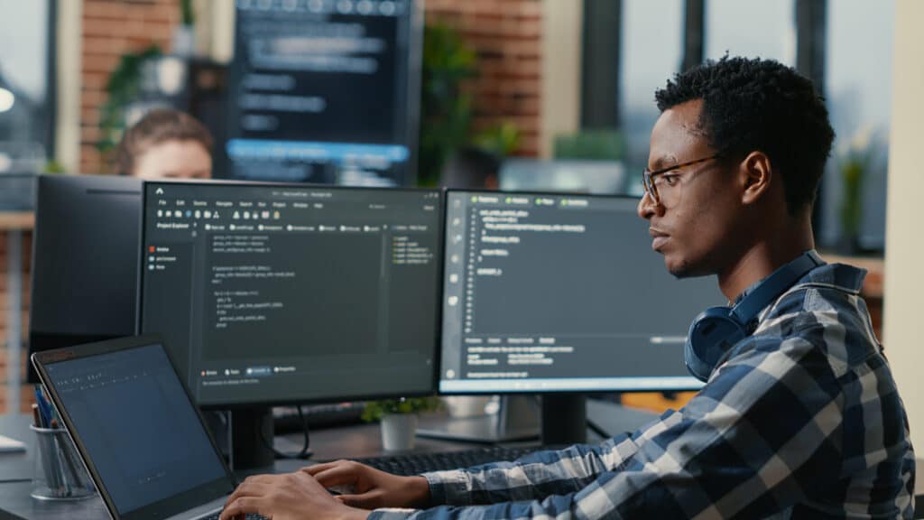A imagem mostra um programador ou analista de segurança cibernética trabalhando em um ambiente moderno de tecnologia. Ele está concentrado em duas telas com códigos abertos e um notebook ao lado, sugerindo atividades de programação, testes ou monitoramento de sistemas. O profissional usa óculos, está com fones de ouvido no pescoço e veste camisa xadrez, reforçando o clima informal típico de ambientes de TI. O cenário, com monitores adicionais ao fundo e iluminação natural, sugere um ambiente de colaboração voltado à inovação e cibersegurança.