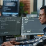 A imagem mostra um programador ou analista de segurança cibernética trabalhando em um ambiente moderno de tecnologia. Ele está concentrado em duas telas com códigos abertos e um notebook ao lado, sugerindo atividades de programação, testes ou monitoramento de sistemas. O profissional usa óculos, está com fones de ouvido no pescoço e veste camisa xadrez, reforçando o clima informal típico de ambientes de TI. O cenário, com monitores adicionais ao fundo e iluminação natural, sugere um ambiente de colaboração voltado à inovação e cibersegurança.