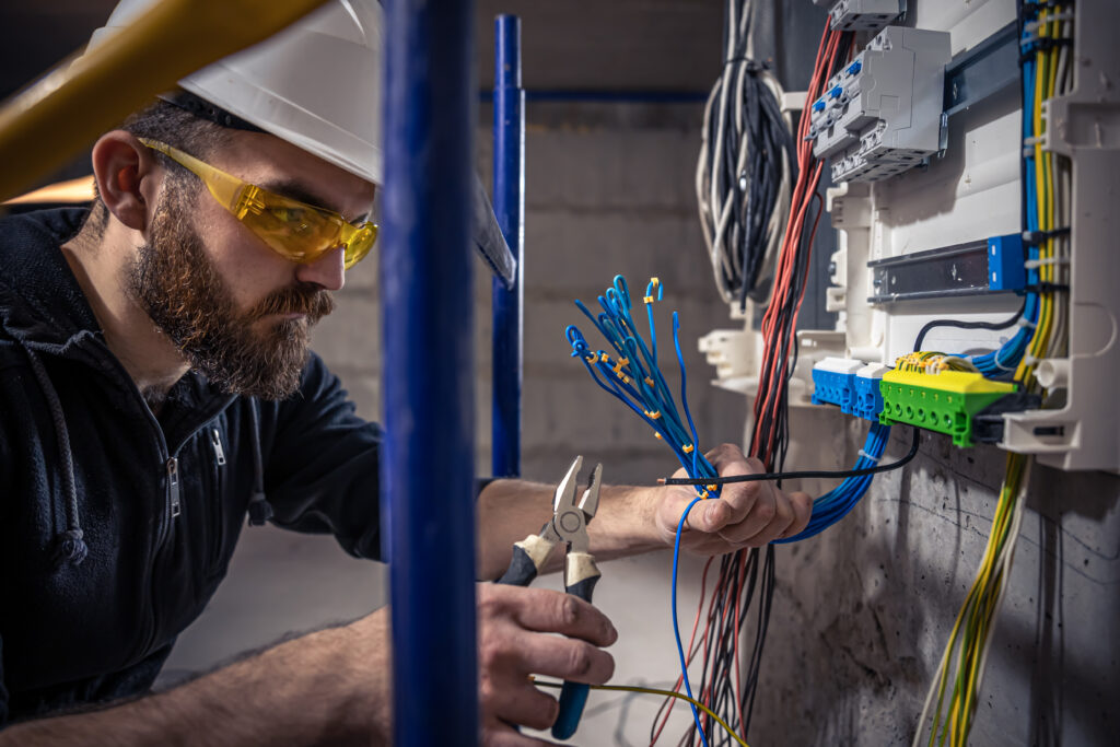 Eletricista com EPI manuseia fios azuis em painel elétrico durante instalação ou manutenção.