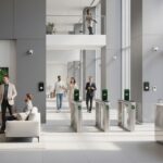 Pessoas em um lobby de escritório moderno, com catracas de segurança e um segurança na recepção. Algumas pessoas estão conversando, enquanto outras estão entrando no edifício.