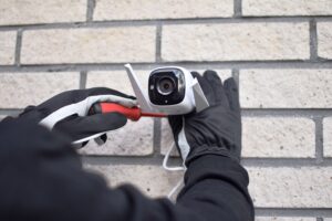 Pessoa tentando desinstalar ou danificar uma câmera de segurança com uma chave de fenda, usando luvas pretas, em uma parede de tijolos.