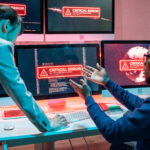 Duas pessoas discutindo sobre um erro crítico em várias telas de computadores, com mensagens de erro vermelho e gráficos de falha nos dispositivos.