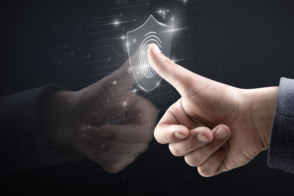 Mão de uma pessoa pressionando o polegar em um ícone holográfico brilhante em formato de escudo com uma digital no centro, simbolizando segurança e autenticação biométrica.