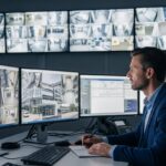 Homem em sala de controle monitorando múltiplas câmeras de segurança em telas grandes e fazendo anotações.