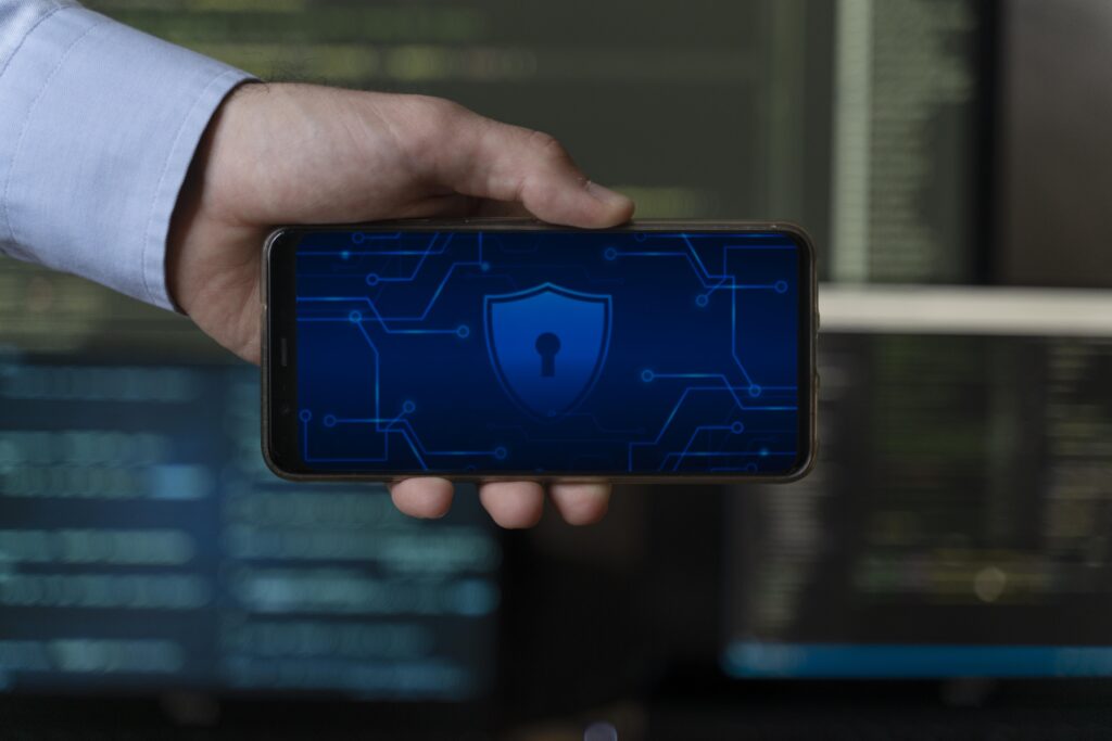 Mão segurando celular com ícone de cadeado e circuito digital sobre fundo de monitores com códigos.