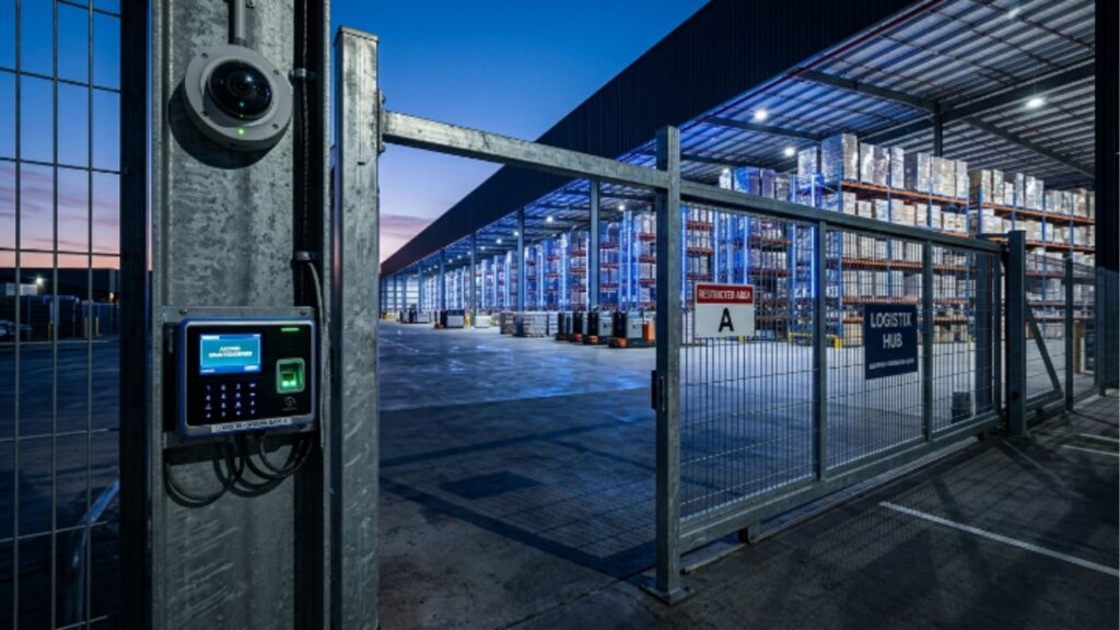 Entrada de um hub logístico ("Logistix Hub") ao anoitecer. Em primeiro plano, um poste de metal sustenta uma câmera de segurança tipo "dome" e um terminal de acesso biométrico com teclado numérico iluminado. Ao fundo, um portão de grade de metal protege o armazém iluminado, onde é possível ver empilhadeiras e fileiras de estoque organizadas. Uma placa no portão indica "Restricted Area A".