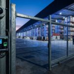 Entrada de um hub logístico ("Logistix Hub") ao anoitecer. Em primeiro plano, um poste de metal sustenta uma câmera de segurança tipo "dome" e um terminal de acesso biométrico com teclado numérico iluminado. Ao fundo, um portão de grade de metal protege o armazém iluminado, onde é possível ver empilhadeiras e fileiras de estoque organizadas. Uma placa no portão indica "Restricted Area A".