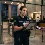 Uma agente de segurança vestindo uniforme preto com a bandeira do Brasil, rádio comunicador e prancheta em mãos. Ela anota informações enquanto inspeciona o crachá de identificação apresentado por um visitante. Ao fundo, veem-se as catracas de acesso e a recepção envidraçada do edifício ao anoitecer.